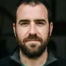 Aurpegian bizarra eta ile motza dituen gizona, kamerari begira aurpegi serioarekin, atzealdea lausotuta.